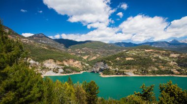 Bir dağ gölünün panoramik manzarası. Temiz turkuaz yeşili su, arka planda bir yol ve çiftçi köyü.