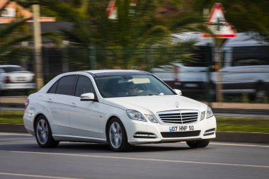 Alanya, Türkiye 12 Nisan 2021: Beyaz Mercedes-Benz E sınıfı sıcak bir yaz gününde palmiyelerin arka planına doğru hızla sürüyor  