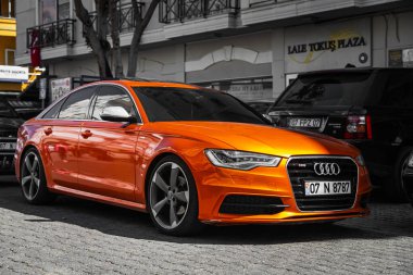 Alanya, Türkiye 17 Nisan 2021: Turuncu Audi A7, sıcak bir yaz gününde bir binanın arka planına, ağaçlara park etti. 