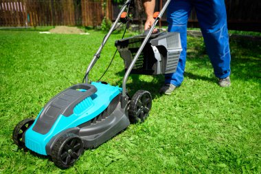 Sokaktaki erkek işçi bir kır evinin yakınındaki modern bir çim biçme makinesinin yardımıyla çimleri biçiyor.