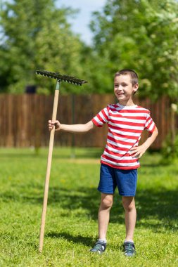 Küçük neşeli bir çocuk elinde tırmıkla bir kır evinin bahçesinde çalışmaya hazır bekliyor..