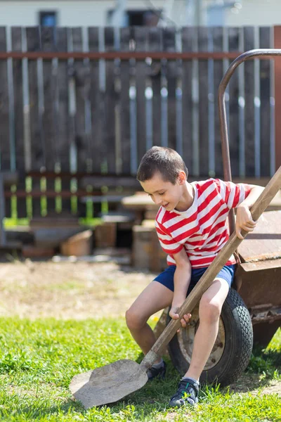 小さな陽気な男の子が庭の車列に座って 田舎の家の庭に彼の手の中にシャベルを持っています 掘る準備ができている小さな男の子のヘルパー — ストック写真