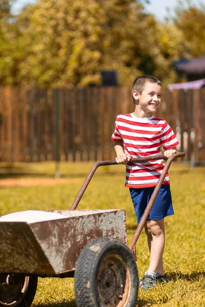 若い笑顔の男の子が庭の周りに車椅子を押しています 田舎でバローと園芸を押して楽しんでいるTショートパンツのボーイヘルパー — ストック写真