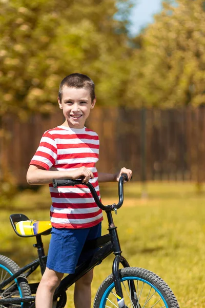 縞模様のTシャツを着た幸せな陽気な少年は カメラを見て 庭の背景に自転車に座って うまくいっています 夏の子供のためのアクティブな屋外ゲーム — ストック写真