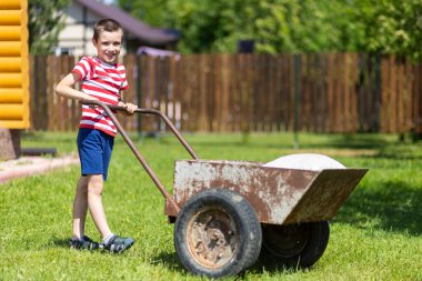 Bahçenin arka bahçesinde el arabasıyla oynayan tatlı çocuk. Gülümseyen çocuk el arabasını bir avluda itiyor..