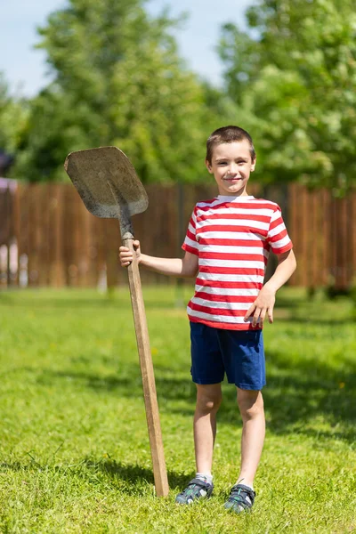 田舎の家の庭で働く準備ができて 彼の手にシャベルを持っている小さな陽気な男の子が立っている — ストック写真