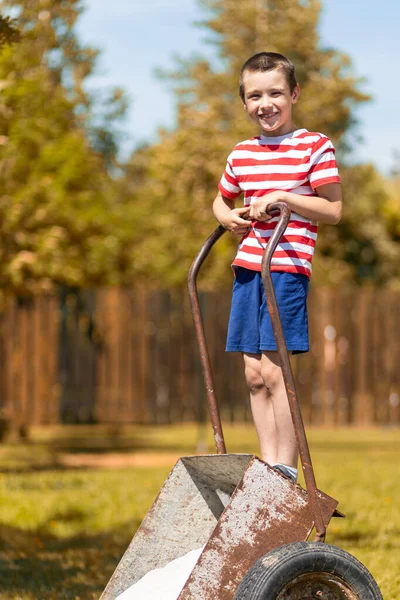 小さな陽気な男の子が庭の車列と田舎の家の庭に立っています 仕事の準備ができている小さなヘルパー少年 — ストック写真