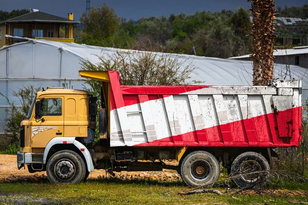 Dump truck with the image of the national flag of Austria is parked against the background of the countryside. The concept of export-import, transportation, national delivery of goods