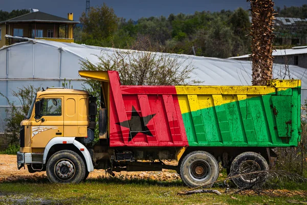 Dump truck with the image of the national flag of Guinea Bissau is parked against the background of the countryside. The concept of export-import, transportation, national delivery of goods