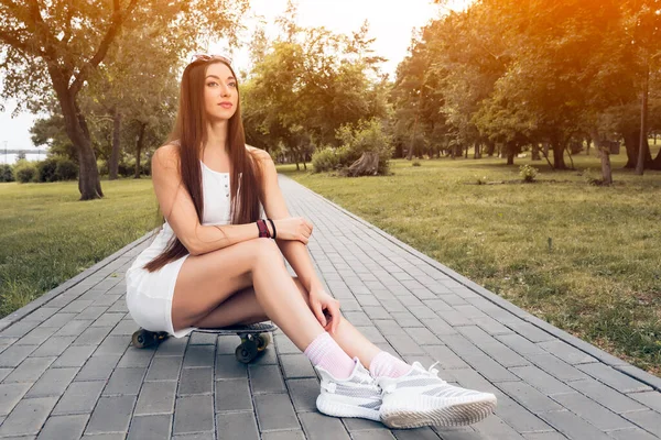 Young athletic woman in sporty style sits on a skateboard  in a city park on a warm summer day. The concept of recreation and walking in an urban environment