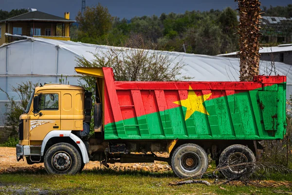 Dump truck with the image of the national flag of Burkino Faso is parked against the background of the countryside. The concept of export-import, transportation, national delivery of goods