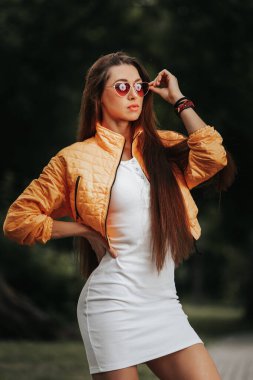 İnce vücutlu güzel, koyu saçlı kadın yeşil şehir parkına karşı etkili bir şekilde poz veriyor.