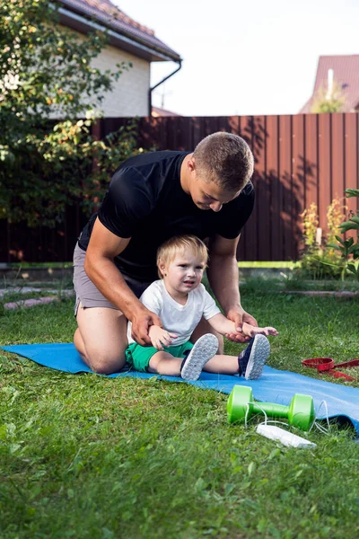 家の近くの庭で暖かい日にマットの上にストレッチ スポーツのために行く彼の小さな陽気な息子と若いお父さんの選手 — ストック写真