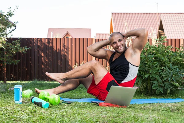 Young man goes in for sports  at home in  backyard in summer day, training online. The athlete makes the press on the mat  in garden