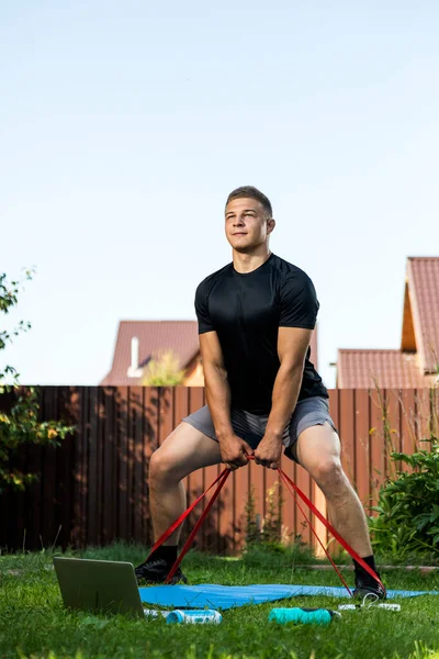 The  strong man goes in for sports in park. Cheerful sporty man with blonde  hair  doing a squat   with rubber and watches  in laptop  in the garden