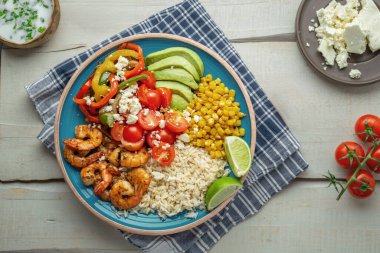 Izgara karides, pirinç, tatlı mısır, kiraz domatesleri, dolmalık biber ve avokado içeren sağlıklı protein dolu bir kase, açık ahşap bir masada sade mavi bir tabakta sunuluyor. Yukardan düz atış.