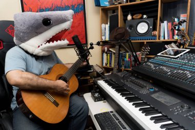 Köpekbalığı kafası takmış bir adam stüdyoda gitar çalıyor.