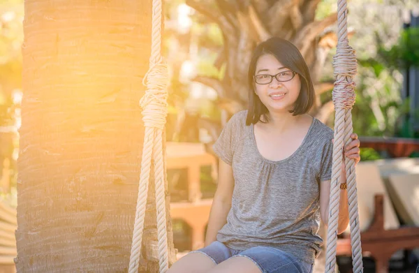 Asian tourist woman, middle aged woman is sitting on wooden swing under coconut or palm tree at resort of Thailand. Wearing eyeglasses, smiling, looking at camera, warm tone image with natural light.