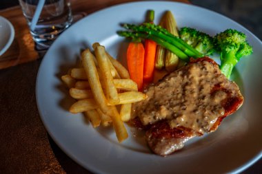 Yuvarlak beyaz tabakta güzel siyah biberli biftek, kızarmış patates, bebek havuç, brokoli, kuşkonmaz ve bebek mısırı. Kara yemek görüntüsü, ahşap masadaki siyah biberli biftek menüsüne yakın çekim..