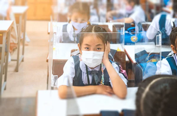 Asian primary school in uniform sitting in classroom, wearing medical mask while studying, plastic shield on student desk, protection student in classroom from covid-19 or corona virus in Thailand.