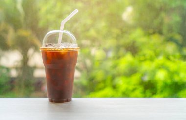 Buzlu Americano kahvesi temizlenmiş plastik bardakta ve beyaz pipetle masaya konuyor, kahve bardağı masanın üstüne konuyor, ağacın arka planı bulanık, sıcak ışık, camın önünde damgalama ve kopyalama için boş alan bırakılıyor..