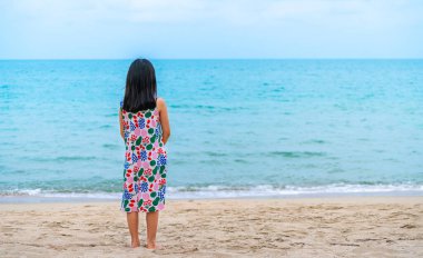 Güzel bir sahilde tek başına duran ve ufka bakan, uzun siyah saçlı, yalınayak, mesaj atmak için boş alan olan Asyalı küçük bir kız çocuğu..
