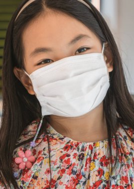 Medikal beyaz maske takan Asyalı şirin bir kızın portresi. Gözler doğrudan kameraya bakıyor..