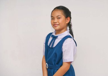 Mavi okul üniformalı Asyalı bir çocuk, sade bir arka plana güvenerek gülümsüyor. Resim eğitim, gençlik ve pozitifliği tasvir ediyor, öğrenme ve okul hayatıyla ilgili konular için ideal.