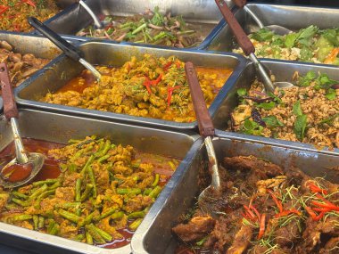 Assorted Thai dishes in stainless steel trays, featuring spicy curries with herbs, vegetables, and meats, served with spoons for self-serve, vibrant, flavorful, and colorful presentation.