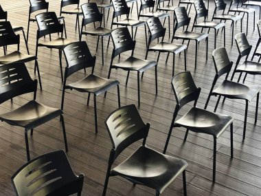 Boş siyah sandalyeler, geniş ve aydınlık bir odada, ahşap bir zeminde dizilmiş, çizgiler ve gölgeler oluşturarak, bir olaya ya da buluşmaya hazırlık hissi uyandırıyor.