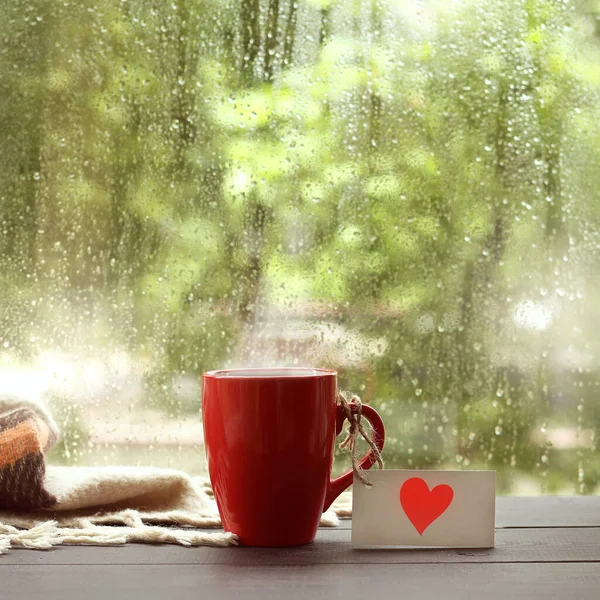 red mug with a note depicting a heart, on the table by the window with drops after rain. warming drink with a love message