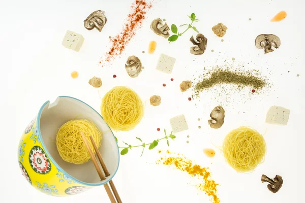 Nidos secos de fideos con otros ingredientes derramados fuera del tazón, aislados sobre fondo blanco . — Foto de Stock