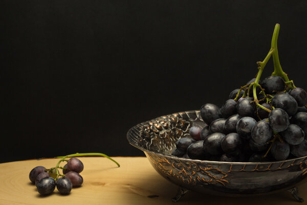 Антикварная чаша с виноградом, на деревянной поверхности и черном фоне
 
