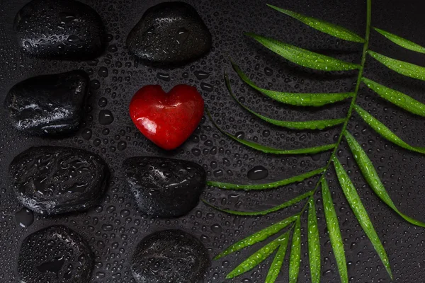 Pierre humide en forme de coeur rouge avec des pierres de basalte noires et des feuilles vertes, sur fond noir avec des gouttelettes d'eau — Photo