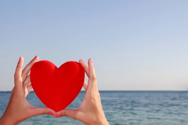 Mani che tengono il cuore rosso con cielo blu e orizzonte marino sullo sfondo . — Foto Stock