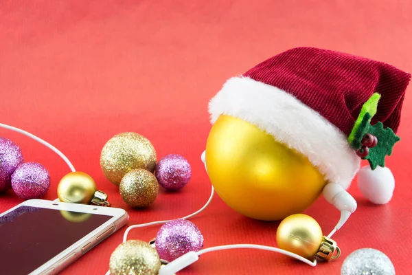 Christmas ball with Santa's hat and smartphone with earphones, on red with copy-space — Stock Photo, Image