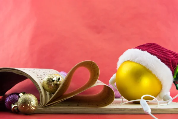 Bola de Natal com chapéu de Papai Noel e fones de ouvido, livro de notação musical com páginas moldando o coração, no fundo vermelho . — Fotografia de Stock