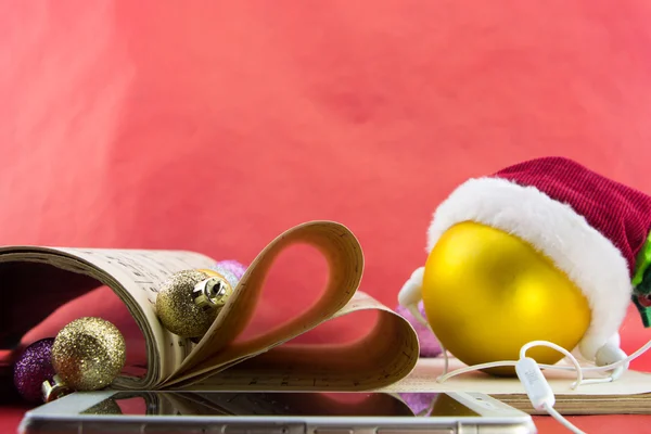 Palla di Natale con cappello e auricolari di Babbo Natale, taccuino musicale con pagine che plasmano il cuore, su sfondo rosso . — Foto Stock