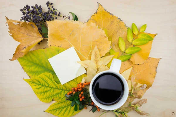 Une tasse de café et une note avec bouquet de feuilles d'automne jaunes et vertes, de baies et de graines . — Photo