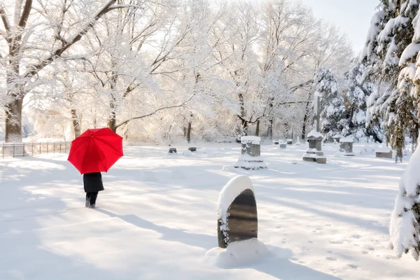 Une personne avec un parapluie rouge dans un cimetière en hiver avec de la neige au sol . — Photo