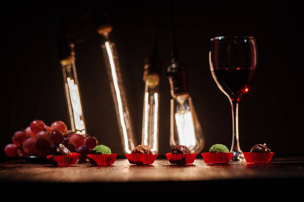 six chocolates in red wrappers on a dark wooden table against the background of a glass with red wine grapes and edison lamps