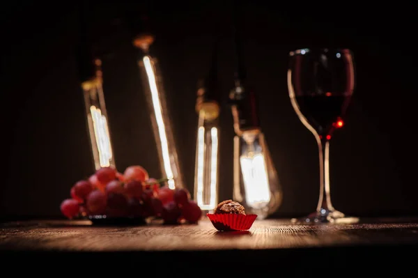 chocolate candy in a red wrapper on a dark wooden table against the background of a glass with red wine grapes and edison lamps