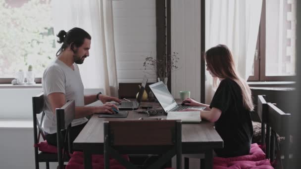 一个年轻的男人和一个年轻的女孩正在他们明亮的公寓里的笔记本电脑前工作 检疫期间在家工作 自由职业者的远程工作 漂亮的年轻沉思女孩 戴着眼镜 站在电脑显示器前 — 图库视频影像