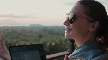 Serbest çalışan kadın yüksek bar taburesinde dizüstü bilgisayarla oturuyor. İnanılmaz günbatımı manzarası ve mavi deniz kıyısında sık bir orman. Gözlüklü ve at kuyruklu hoş bir kız bir kafede bilgisayar başında çalışıyor..