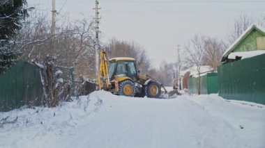 Sarı bir traktör kışın küçük bir köyün caddesinde kar temizler. Kış güneşli gün.