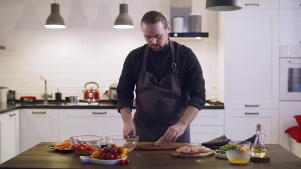 Jonge schattige chef snijdt kippenvlees op een houten plank in de thuiskeuken. — Stockvideo