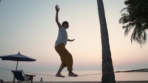 Das Mädchen auf einer Slackline bei Sonnenuntergang an einem tropischen Strand. — Stockvideo