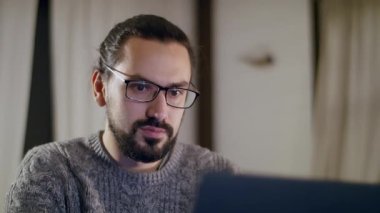 Örgü kazaklı, genç, yakışıklı, beyaz bir adam evde dizüstü bilgisayar kullanarak çalışıyor. Freelancer evden rahat çalışıyor. Akşamları bilgisayar ekranının önünde gözlüklü bir adam..