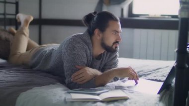 Genç ve yakışıklı bir adam yatakta yatarken dizüstü bilgisayarda çalışıyor..