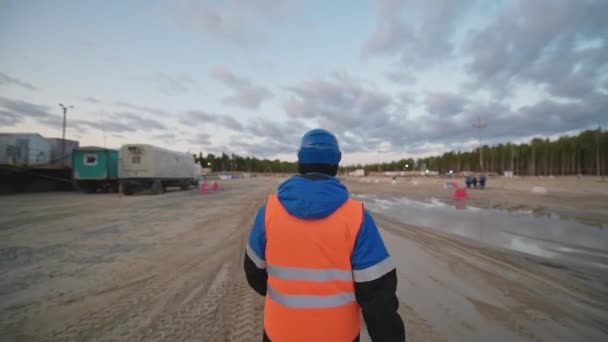 Un uomo con un giubbotto protettivo arancione cammina attraverso un giacimento petrolifero. — Video Stock